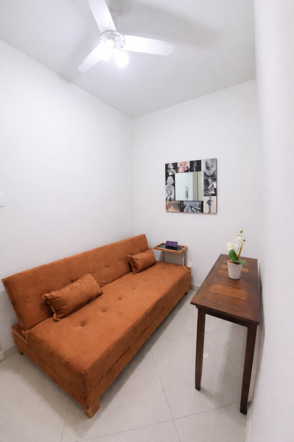 Una habitación pequeña y luminosa con un sofá, una mesita con una planta, un ventilador de techo y un espejo montado en la pared.