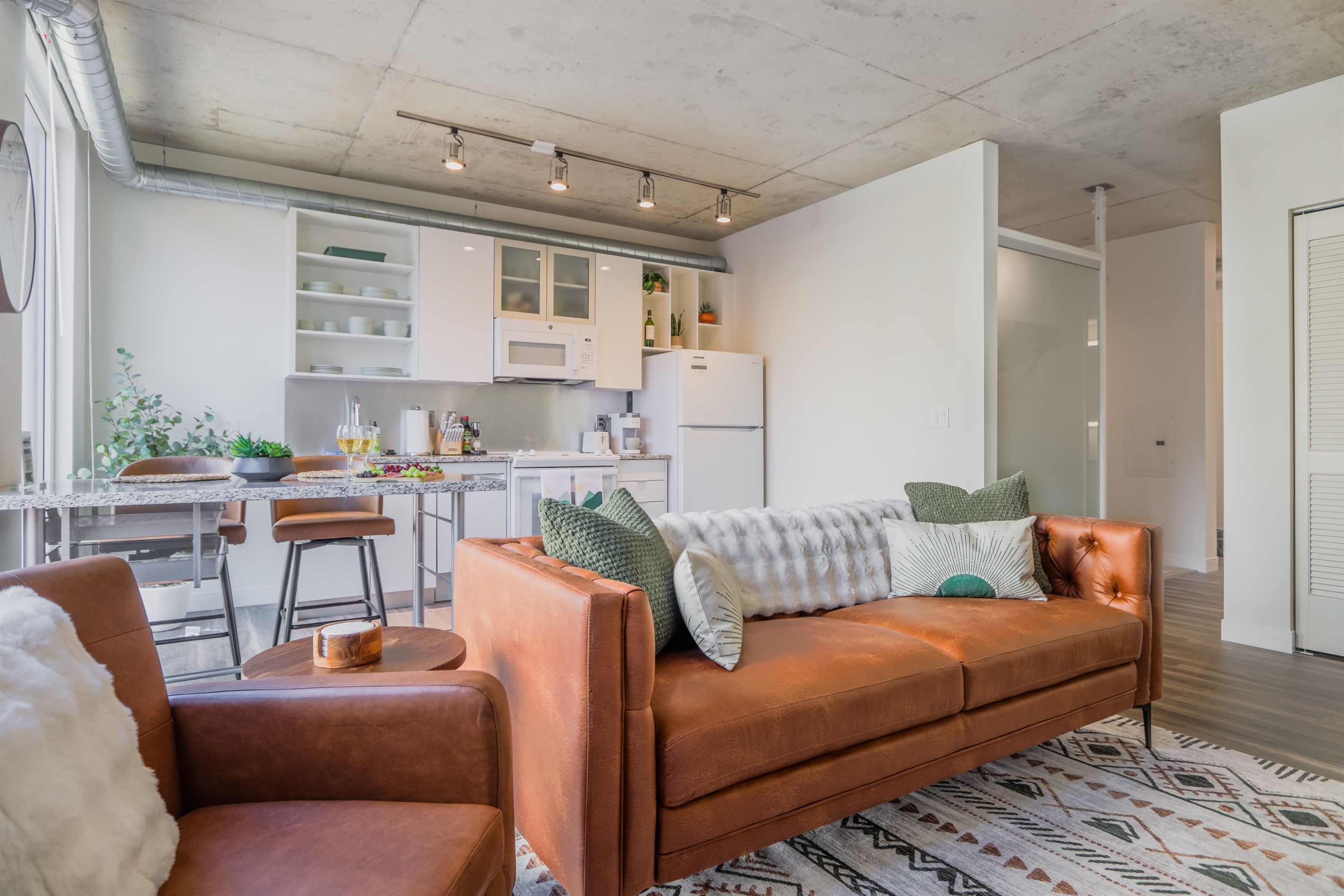 A modern kitchen with white cabinets, a dining area with a table and chairs, and a cozy living space with a brown sofa and cushions.