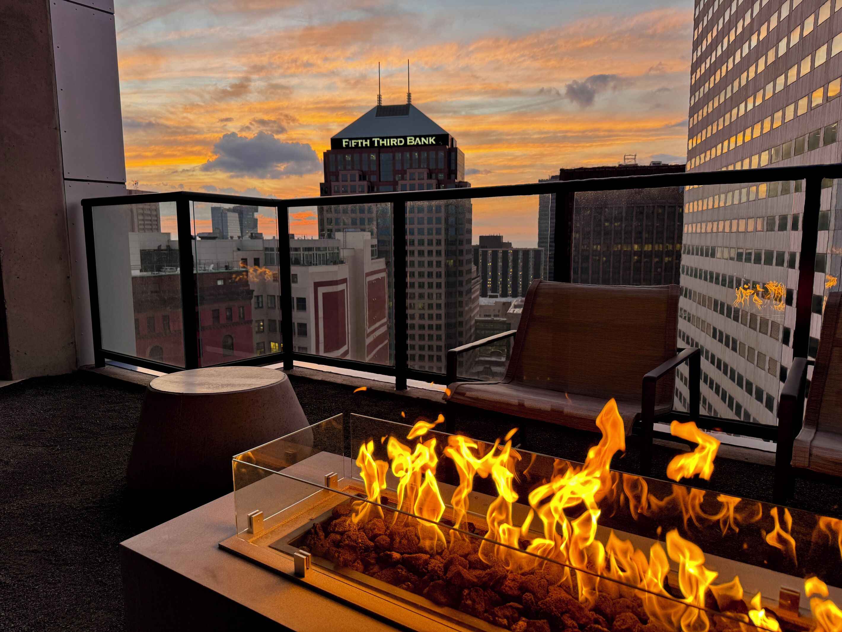 A balcony with a glass railing, a fire feature, and a view of a cityscape including a building labeled 'Fifth Third Bank.'