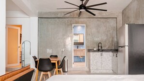 A modern hotel room with a bed, a dining table with chairs, a kitchenette, and a ceiling fan. - Cozy Studio Suite - Art District (San Juan)