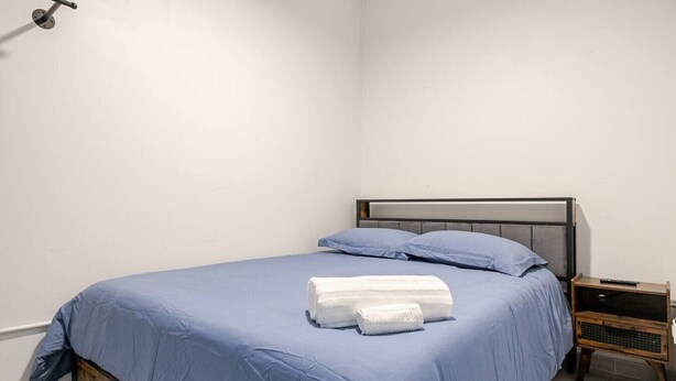 A neatly made bed with blue bedding and white towels, a wooden nightstand with a remote control, and a plain white wall. - Cozy Studio Suite - Art District (San Juan)