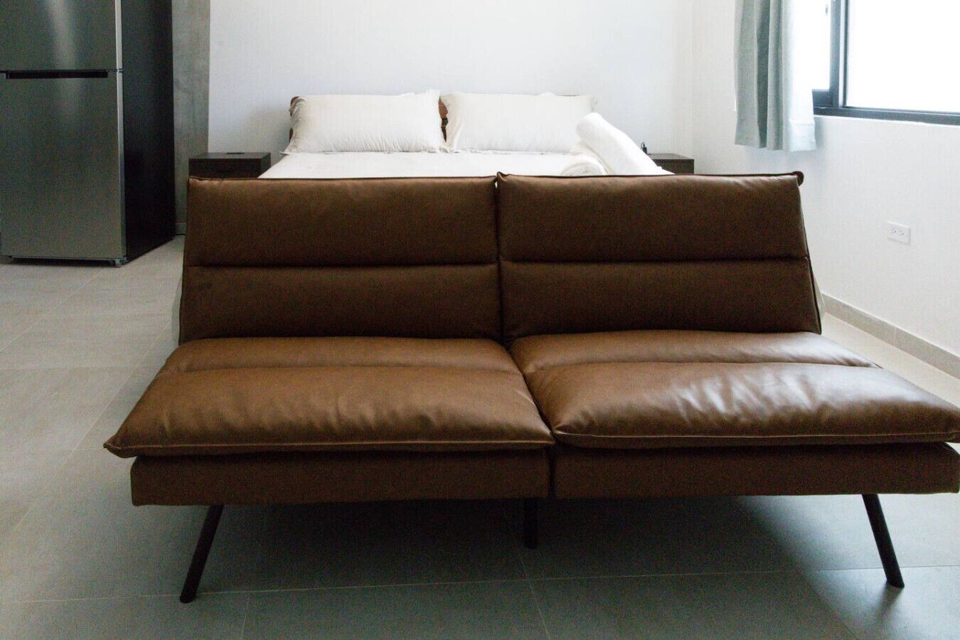 Una habitación moderna con un sofá de cuero marrón, una cama con ropa blanca y un ventanal con cortinas.