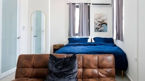 A bedroom with a bed, a brown leather sofa with a black pillow, a glass shower door, and a painting on the wall. - Modern 2 Bed Room Suite in Art District (San Juan)