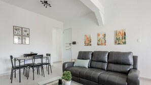 A modern living room with a black leather sofa, a glass coffee table, a dining area with a black metal table and chairs, and a door leading to another room. - Modern 2 Bed Room Suite in Art District (San Juan)