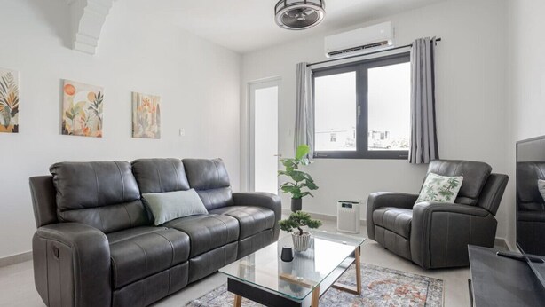 A modern living room with a leather sofa, a glass coffee table, and a grey armchair. There is a large window with curtains, a ceiling fan, and an air conditioning unit. - Modern 2 Bed Room Suite in Art District (San Juan)