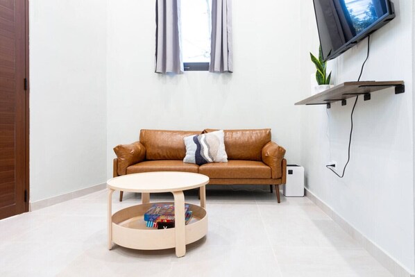 A modern living room with a brown leather sofa, a round wooden coffee table, a flat-screen TV mounted on the wall, and a small potted plant. - Cozy 2 Bed Room in Art District (San Juan)