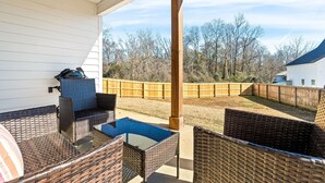 Een overdekt terras met rieten meubilair en een tafel, met uitzicht op een omheinde tuin met bomen.