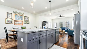 Een moderne keuken met grijze kastjes, een granieten aanrechtblad en een centraal eiland. Er hangen foto’s in lijsten aan de muur, een eethoek met stoelen en een zithoek met een bank en een televisie.