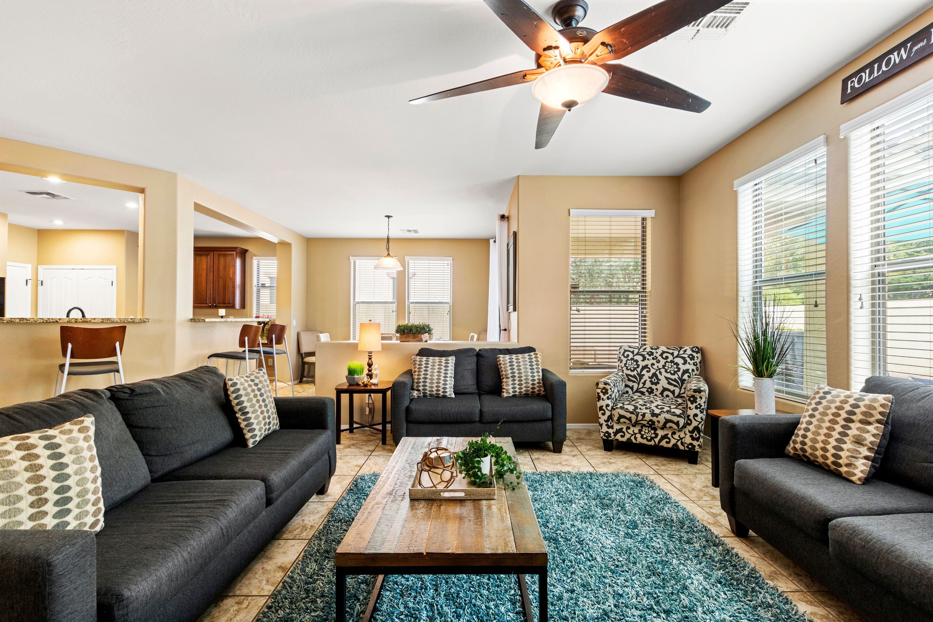 A modern living room with a dark grey sofa set, a wooden coffee table, and patterned throw pillows. A ceiling fan is visible, and the room has large windows with blinds.