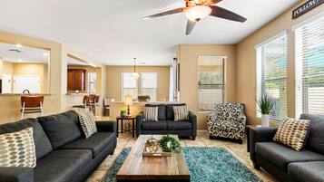 A modern living room with a dark grey sofa set, a wooden coffee table, and patterned throw pillows. A ceiling fan is visible, and the room has large windows with blinds.