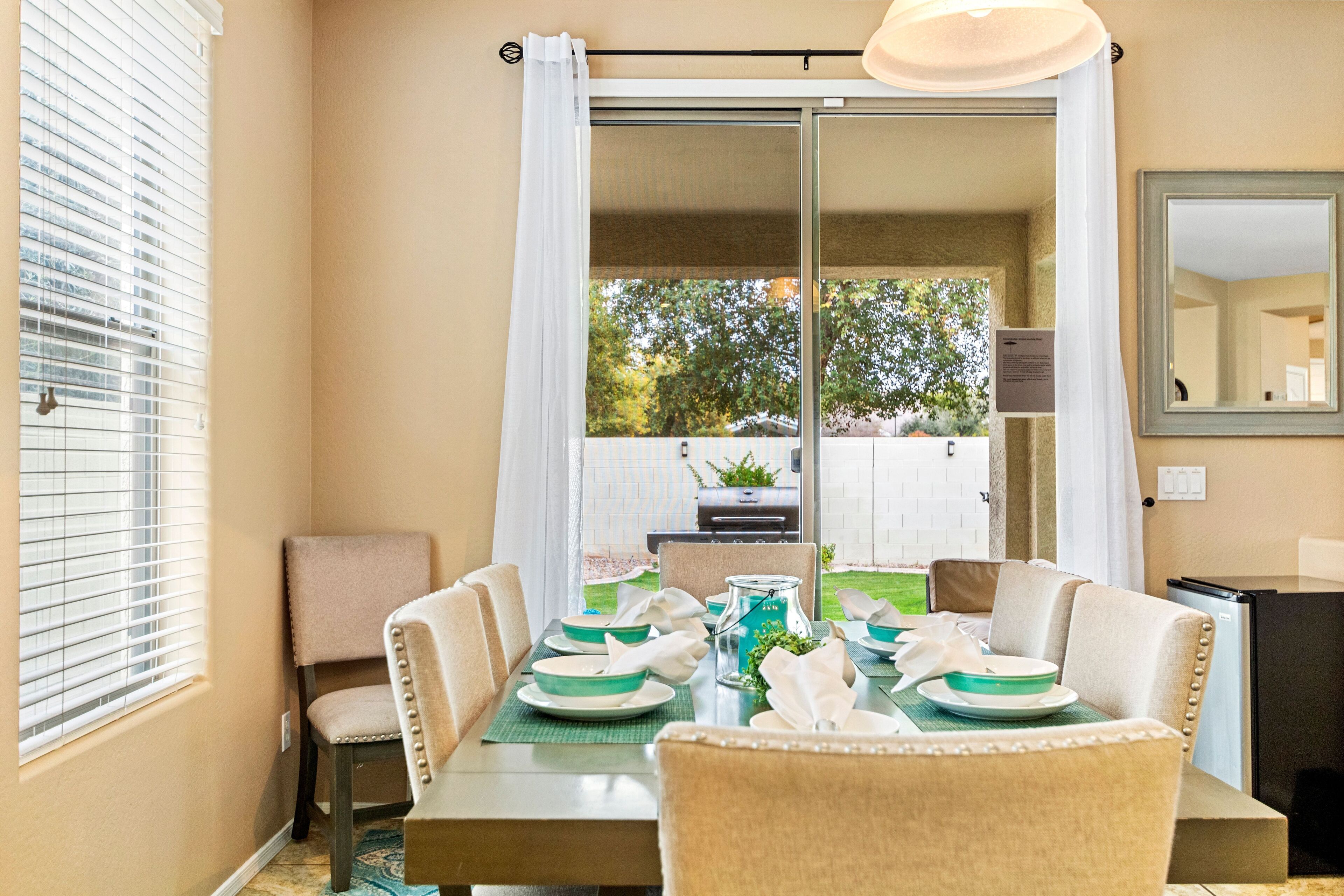 A dining area with a table set for four, a chair, a grill, and a view of the backyard.