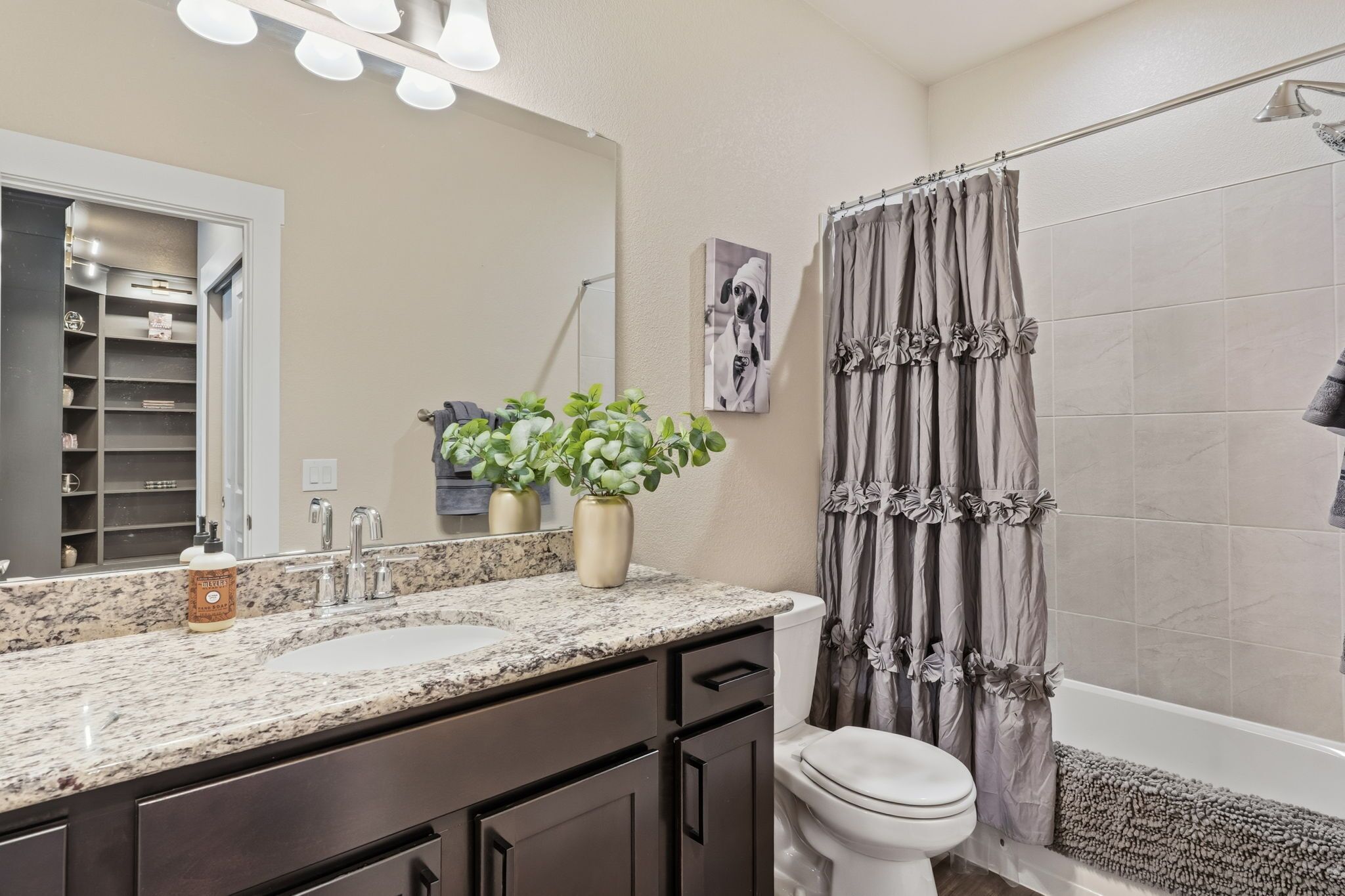 A bathroom with a granite countertop, two sinks, a toilet, a shower with a curtain, and a mirror with lights.