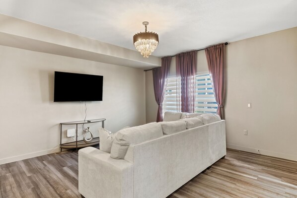 Un salon moderne doté d’un grand téléviseur à écran plat, d’un canapé beige, d’un lustre et d’une fenêtre avec des stores et des rideaux.