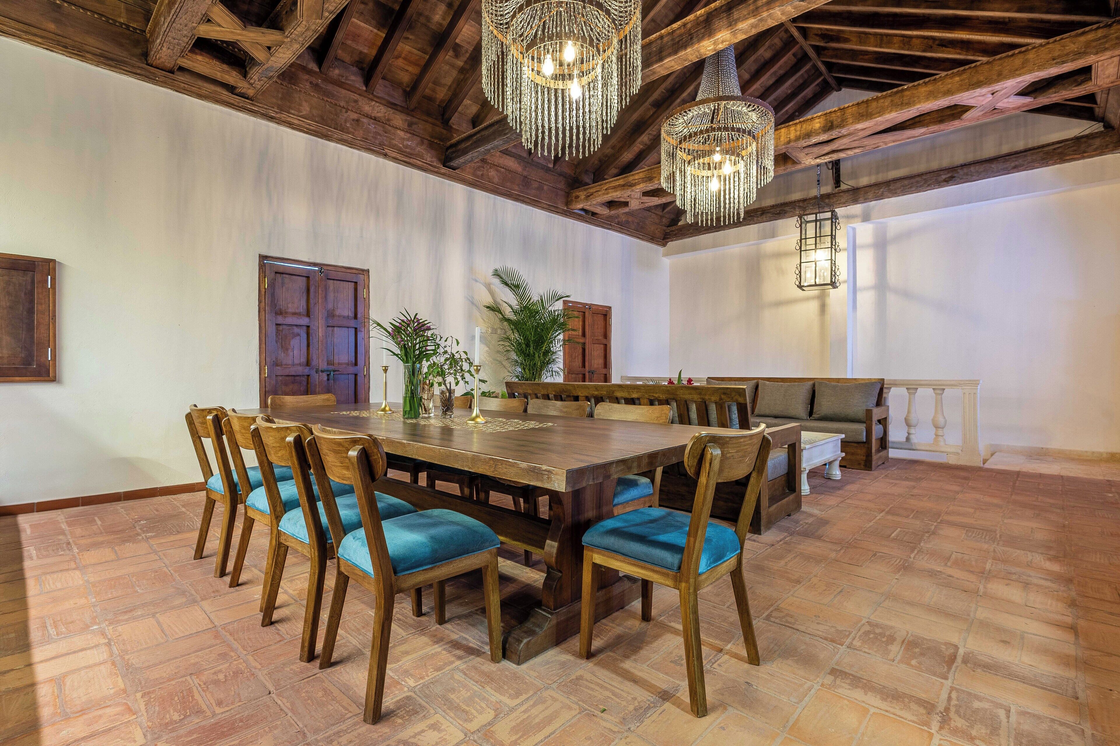 Un comedor amplio con una gran mesa de madera rodeada de sillas con cojines azules, una araña y un techo de madera.