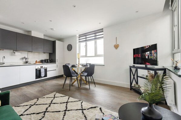 Una cocina moderna con armarios blancos, un comedor con mesa negra y detalles dorados, y un televisor sobre un soporte negro.