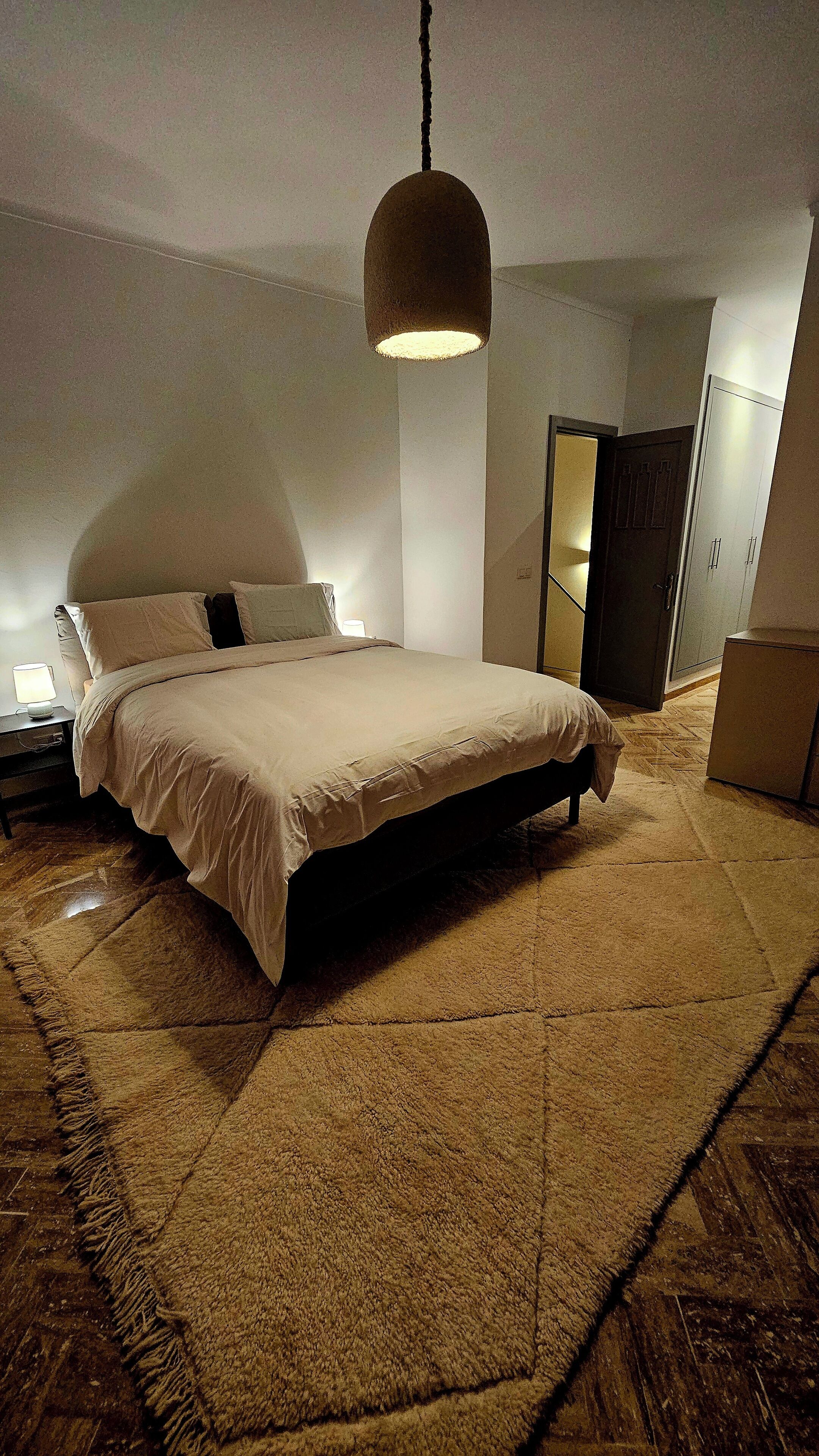 A modern bedroom with a large bed, a hanging light fixture, a wardrobe, and a mirror.