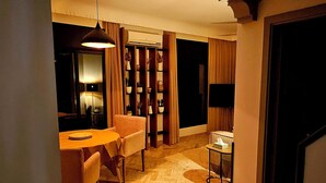 A warmly lit interior with a wooden floor, a dining area, a sofa, a TV, and a bookshelf.