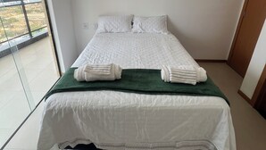 En snyggt bäddad säng med vita lakan och en grön pläd, ett fönster med utsikt över byggnader och en dörr till höger.