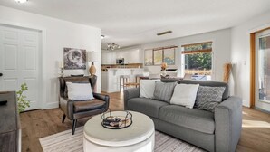 A modern living room with a grey sofa, a leather armchair, and a white coffee table. There is a kitchen in the background with white cabinets and a dining area. - Bighorn Manor | Central | Wood Fireplace | Views (Radium Hot Springs)