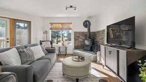A modern living room with a stone fireplace, a sofa, a television, and a view of the outdoors. - Bighorn Manor | Central | Wood Fireplace | Views (Radium Hot Springs)