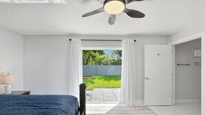 Ein Schlafzimmer mit einem Bett, einem Deckenventilator, einem Nachttisch und Blick auf einen Garten durch eine offene Tür.
