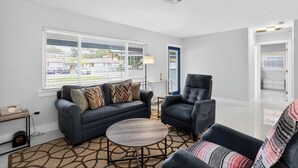 Ein modernes Wohnzimmer mit einem grauen Sofa, einem runden Couchtisch und einem schwarzen Sessel. Im Hintergrund ist ein Fenster mit Jalousien, eine Lampe und eine Tür zu sehen.