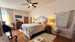 Une chambre à coucher comprenant un lit, des lampes de chevet, un ventilateur de plafond, une commode en bois, une chaise, une petite table avec un livre et une décoration murale à motifs.
