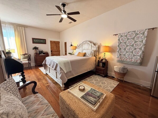 A bedroom with a bed, bedside lamps, a ceiling fan, a wooden dresser, a chair, a small table with a book, and a patterned wall hanging.