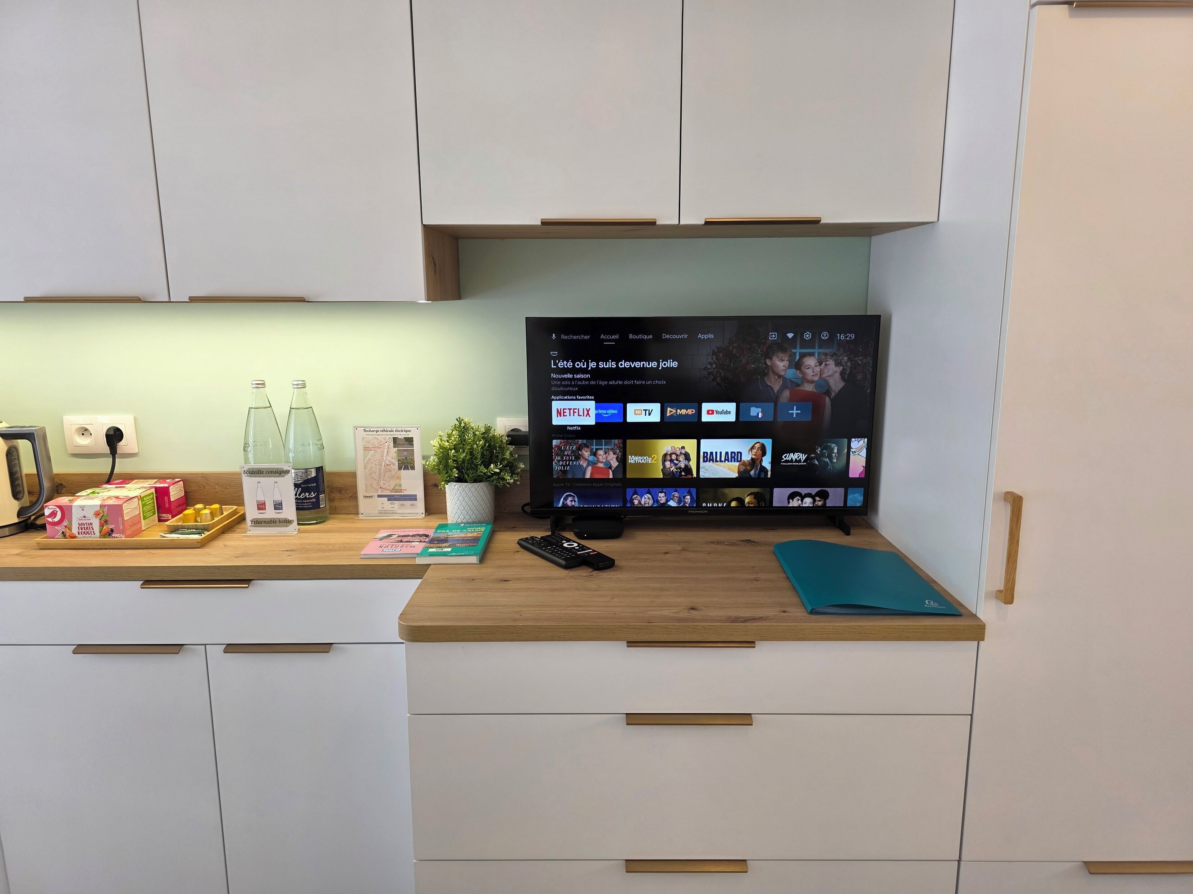 Een keuken met een flatscreen televisie waarop een streamingdienst draait, een waterkoker en diverse keukengerei op het aanrecht.