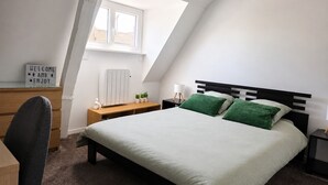 Ein Schlafzimmer mit Bett, Schreibtisch, Stuhl, Heizkörper, Fenster und Nachttisch mit Lampe.