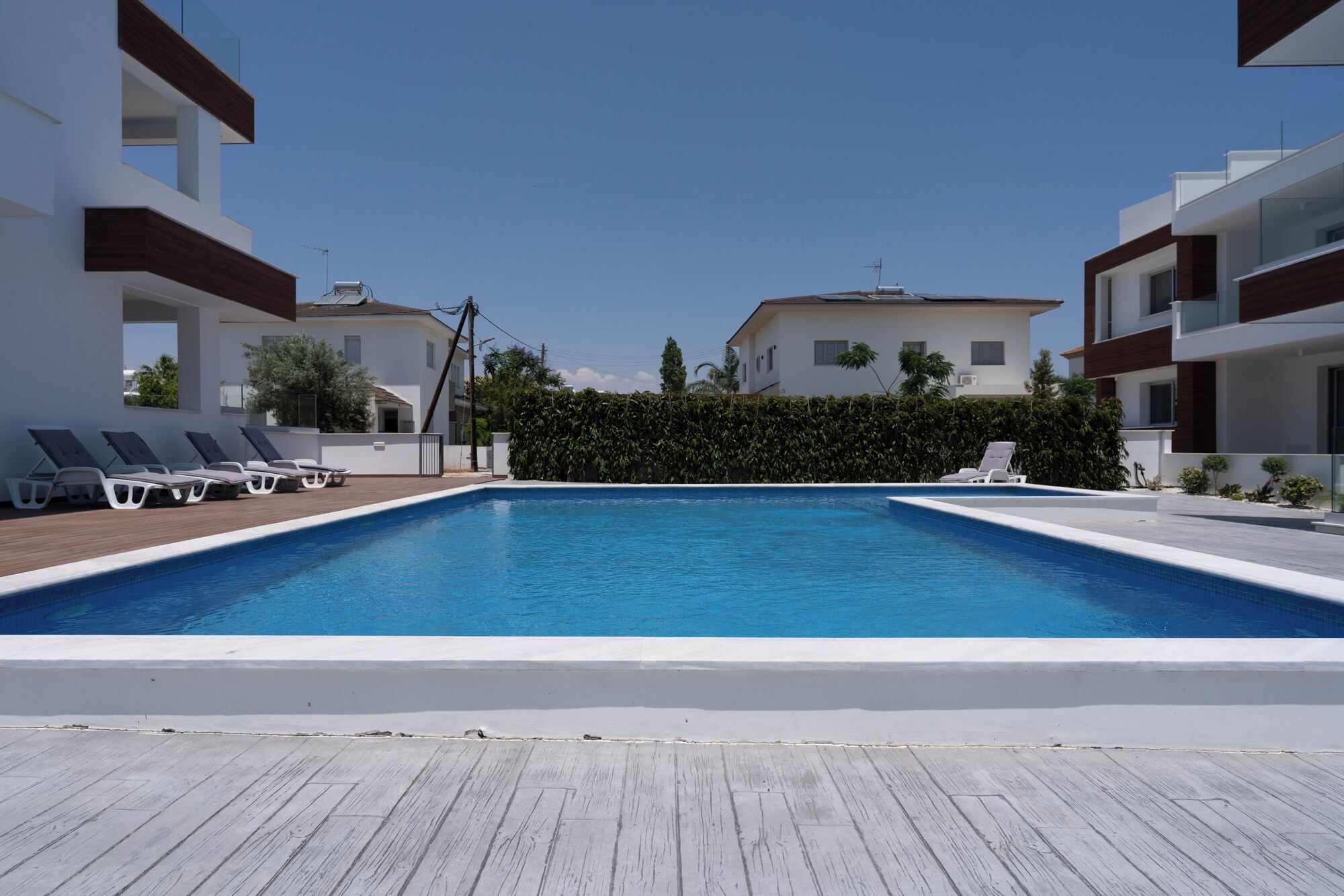 Ein rechteckiger Swimmingpool mit einer Holzdielen-Terrasse im Vordergrund, umgeben von modernen Gebäuden mit Balkonen und einer Hecke im Hintergrund.