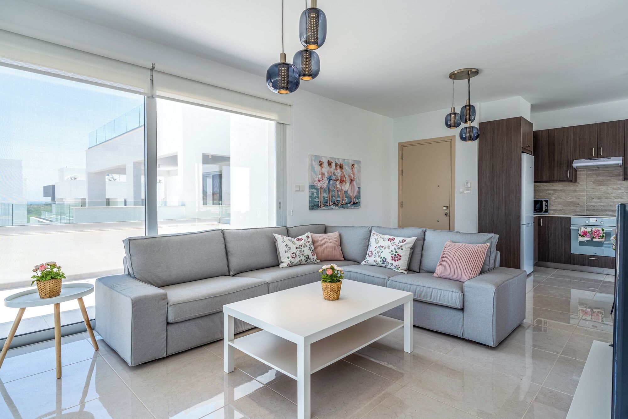 A modern living room with a grey sofa, white coffee table, and decorative pillows. There is a kitchen area with dark wooden cabinets and a white refrigerator.