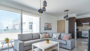 Un saló modern amb un sofà gris, una taula de centre blanca i coixins decoratius. Hi ha una zona de cuina amb armaris de fusta fosca i un frigorífic blanc.