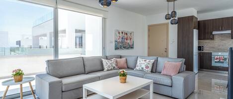 A modern living room with a grey sofa, white coffee table, and decorative pillows. There is a kitchen area with dark wooden cabinets and a white refrigerator.