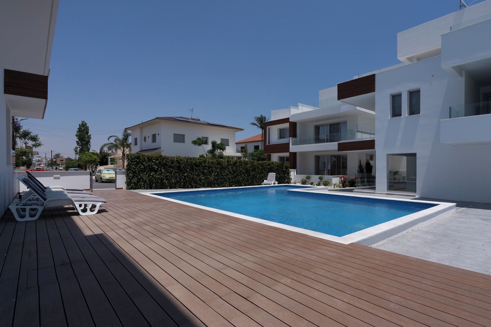 Un área de piscina moderna con una terraza de madera, sillas de descanso y una piscina azul cristalina.
