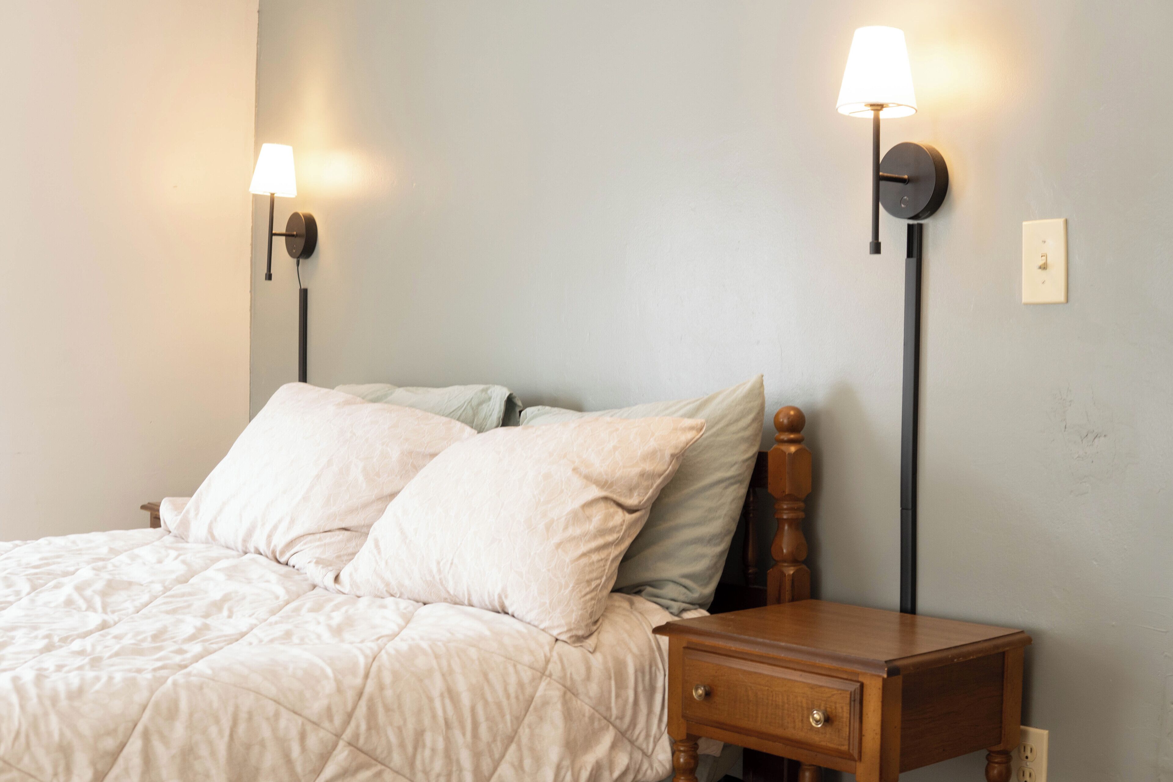 Una cama bien hecha con cubrecama acolchado y dos almohadas, una mesita de noche de madera con cajón, y dos lámparas fijadas a la pared.