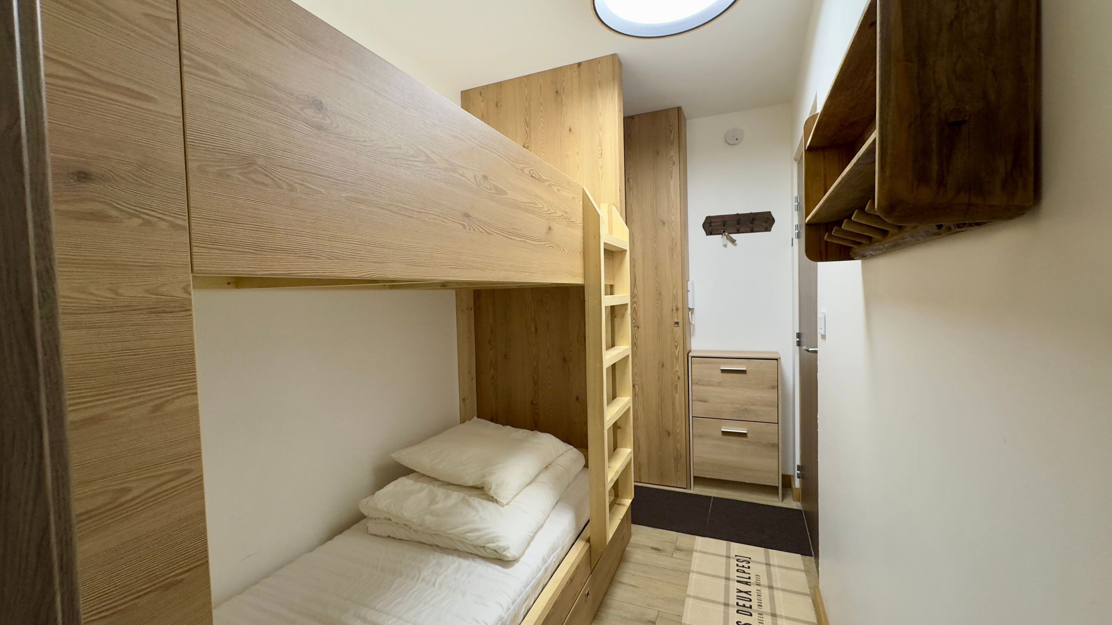 Een kleine kamer met een stapelbed, een houten garderobe en een bureau.