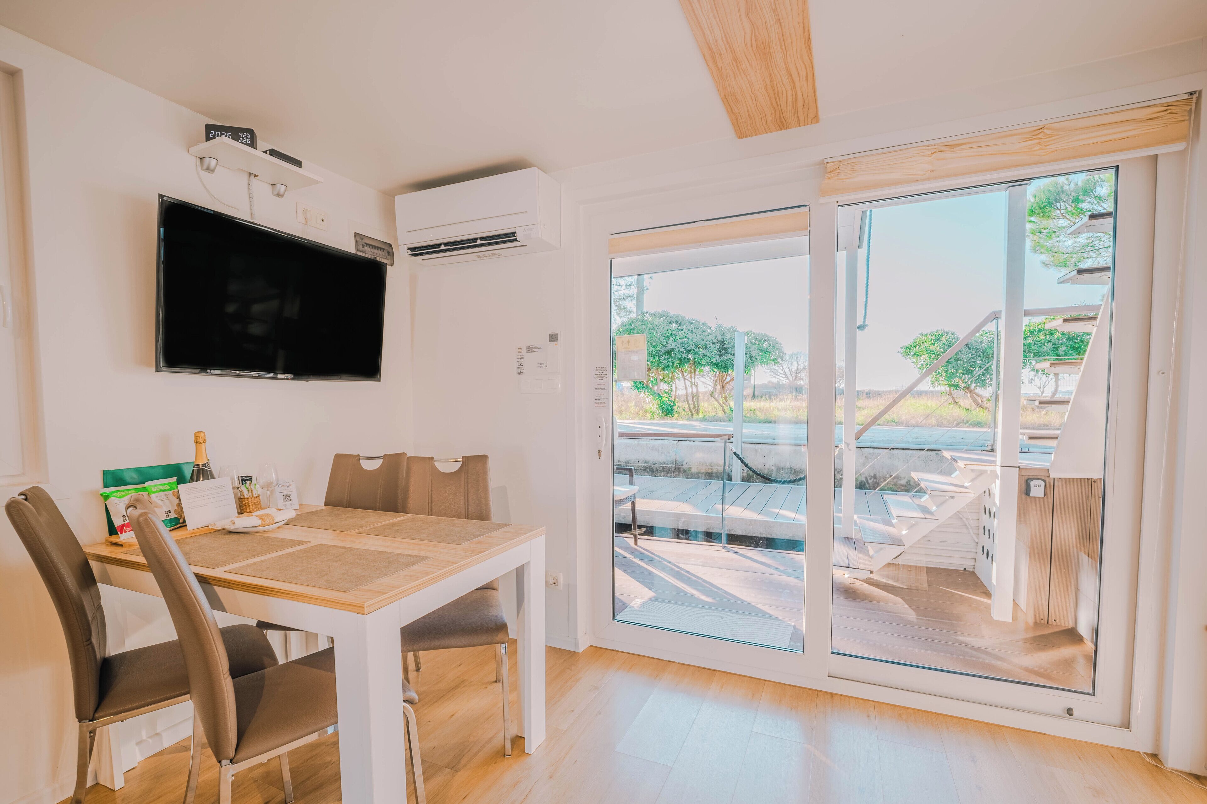 A modern room with a flat-screen TV, a dining table with chairs, a sliding glass door to an outdoor area, and an air conditioning unit.