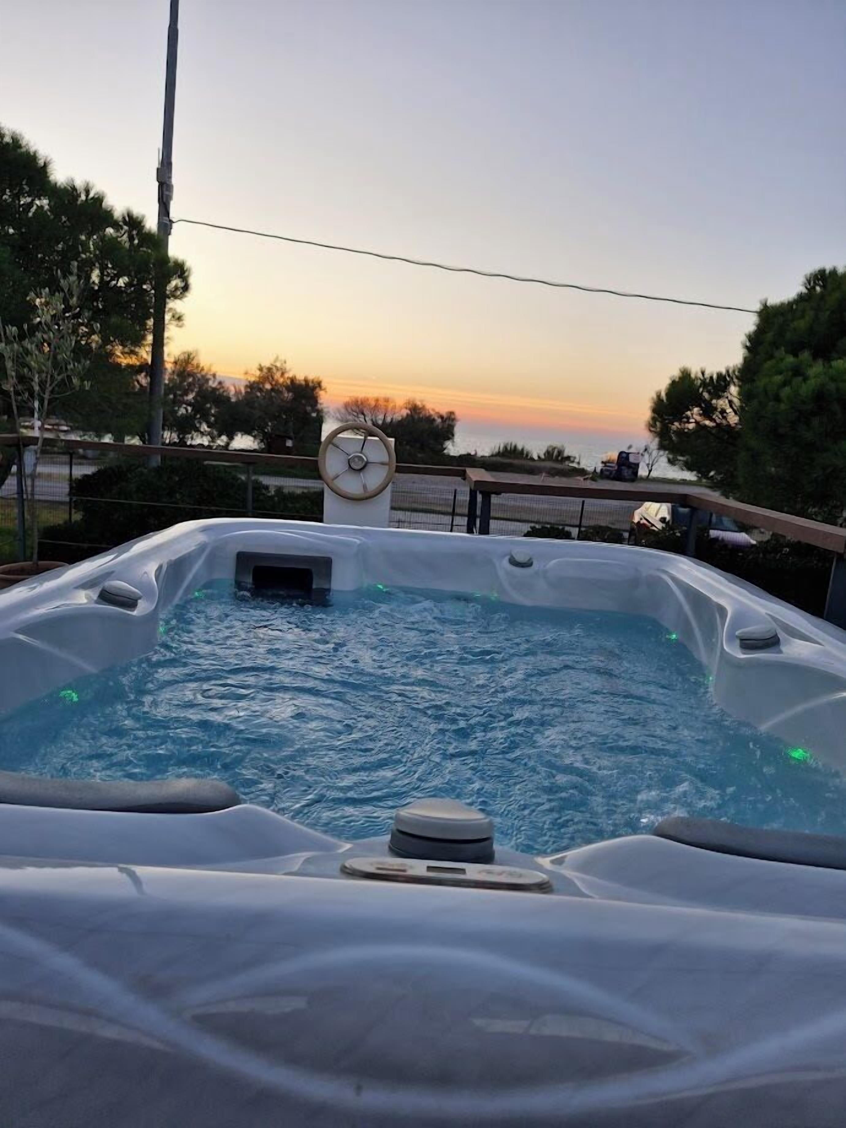 Un jacuzzi con panel de control, rodeado por una terraza y árboles, con un atardecer de fondo.
