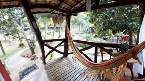 A hammock under a wooden pergola with a view of a garden and a building in the background. - Pousada Brisa Do Vale (Barra do Piraí)