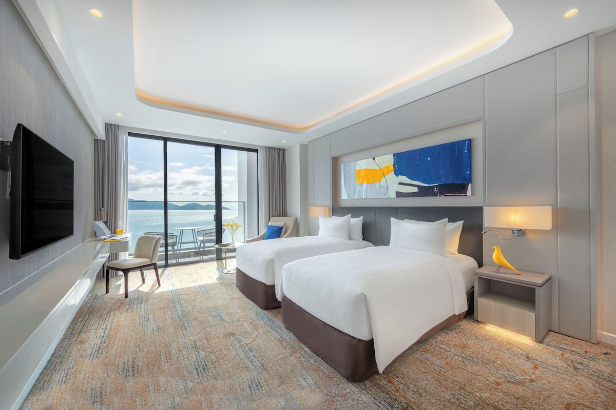 A hotel room with two beds, a TV, a desk, and a view of the sea.