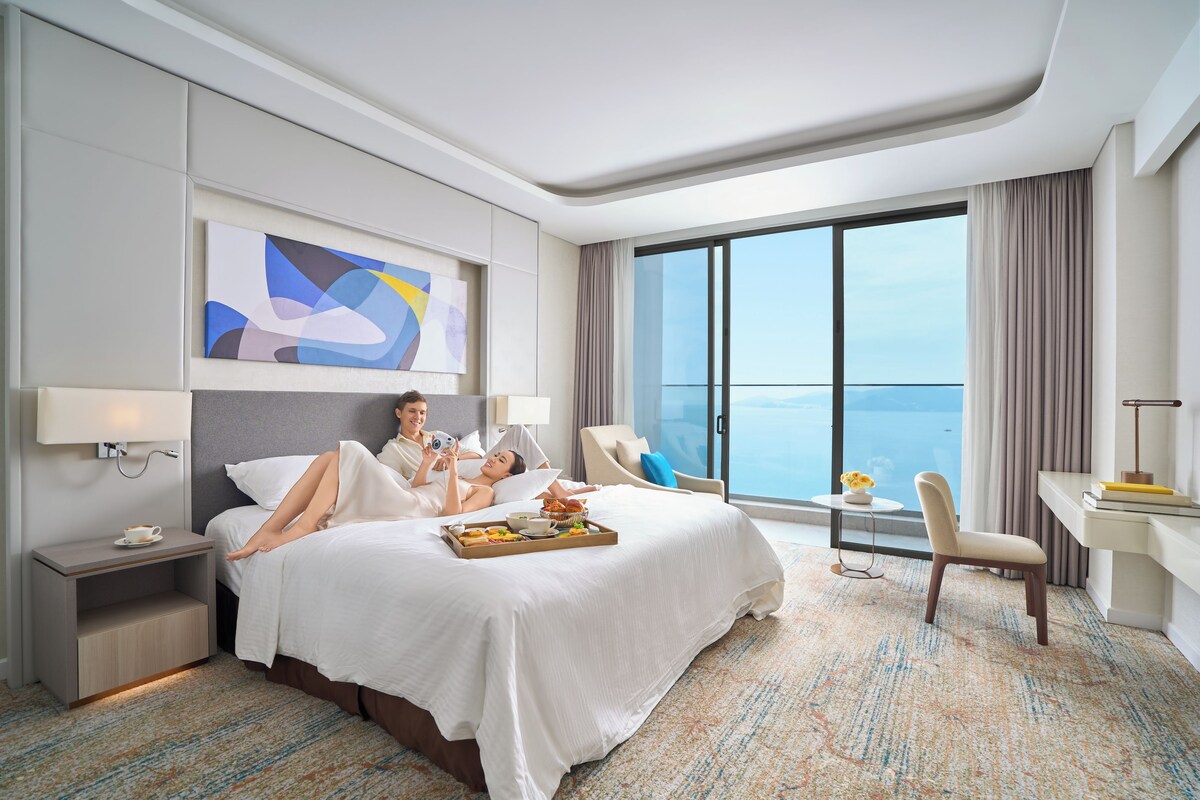 A hotel room with a large bed, a desk, a chair, a view of the ocean, and a painting on the wall.