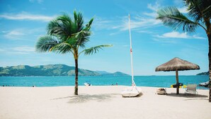 Una playa con un velero, palmeras, una sombrilla de paja y una silla de descanso.