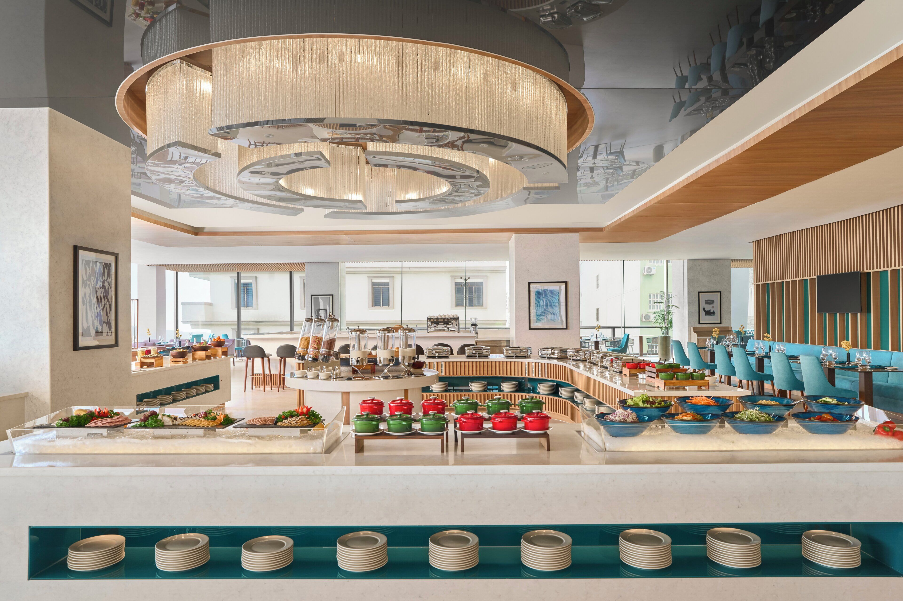 A modern hotel lobby with a central buffet, seating areas, and a large chandelier.