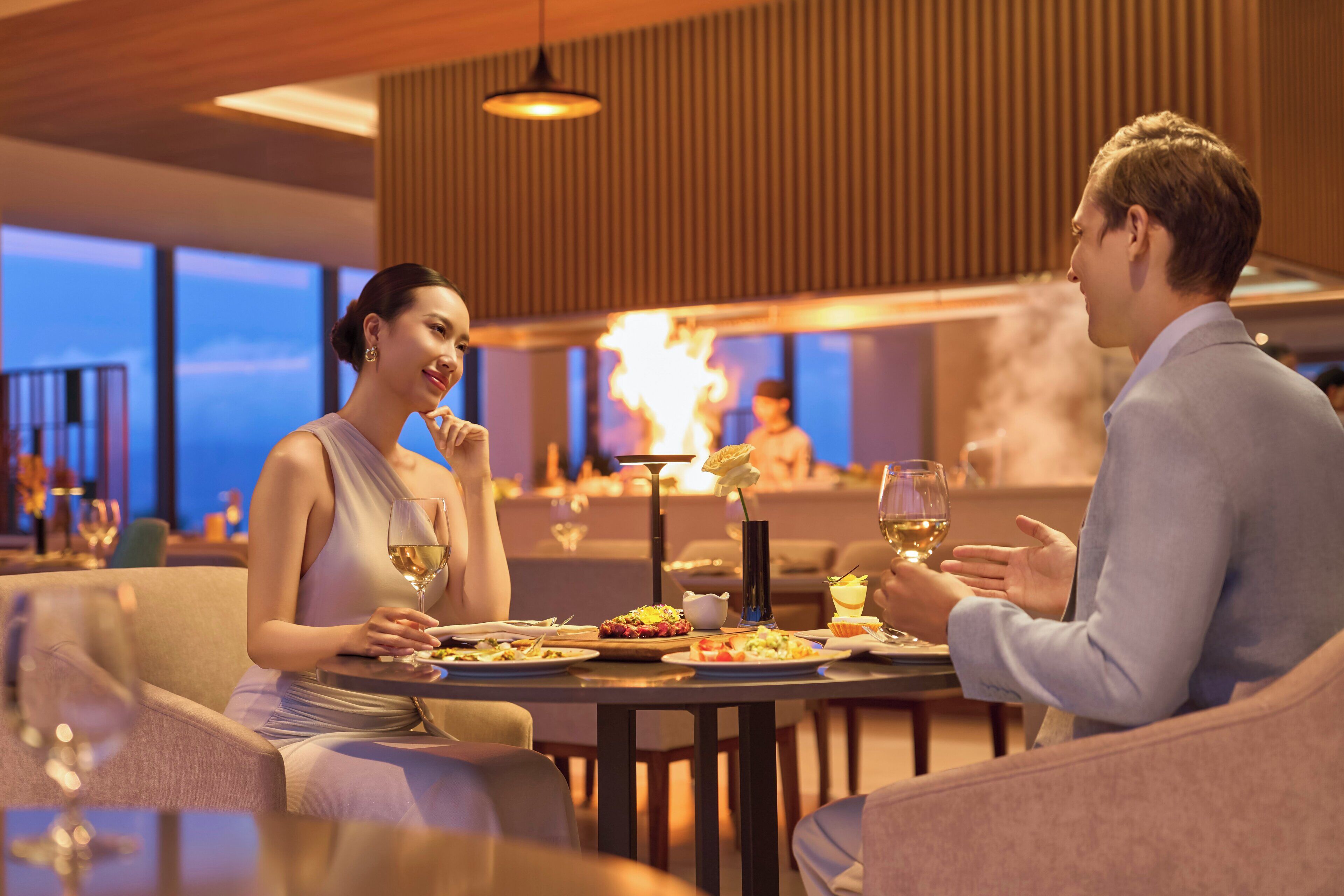 Una pareja cenando en una mesa con vistas a un paisaje urbano y un espectáculo de fuegos artificiales al fondo.