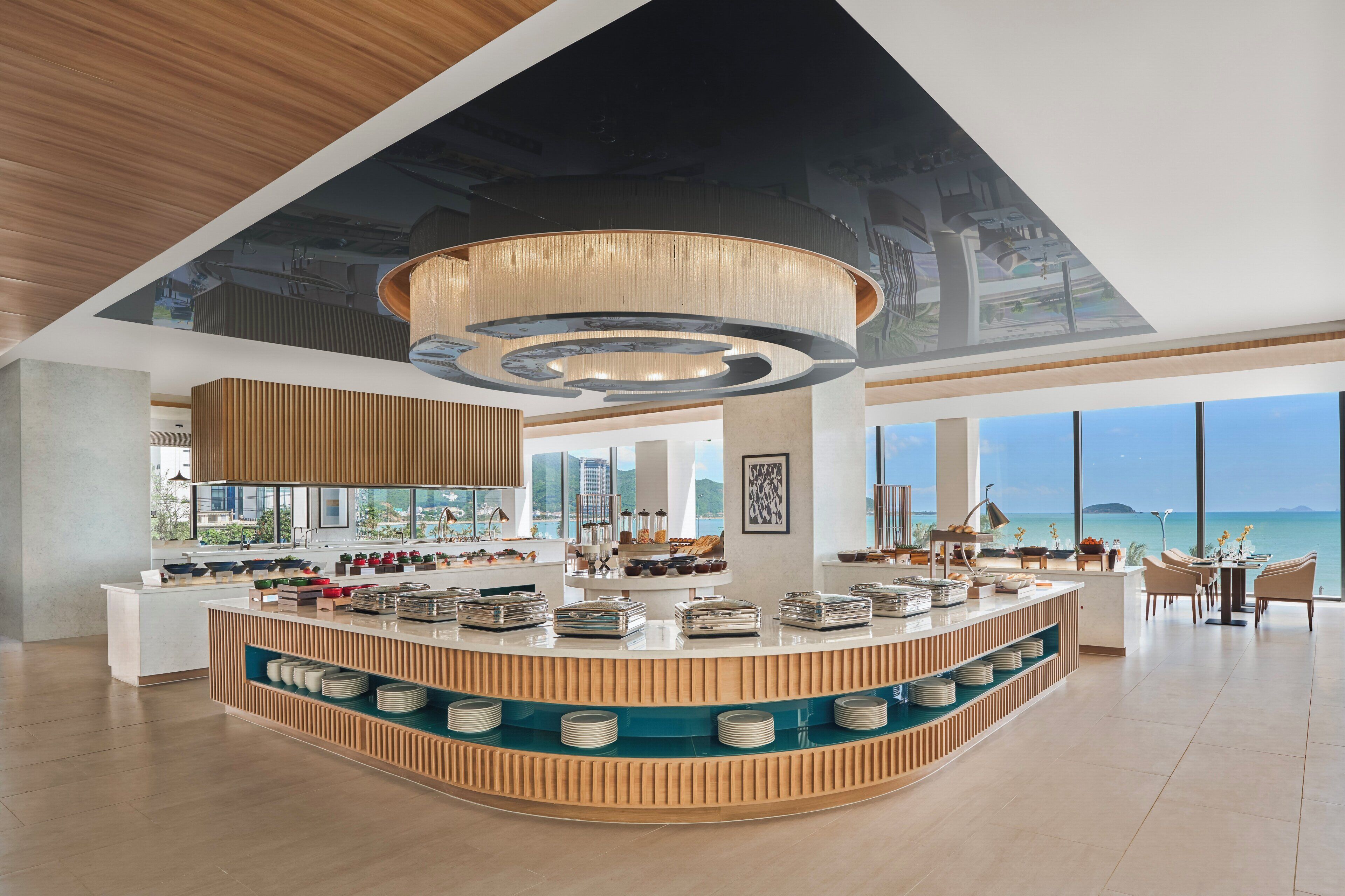Un hall d’hôtel moderne doté d’un comptoir central, de grandes fenêtres offrant une vue sur l’océan et d’un plafond haut orné d’un luminaire original.