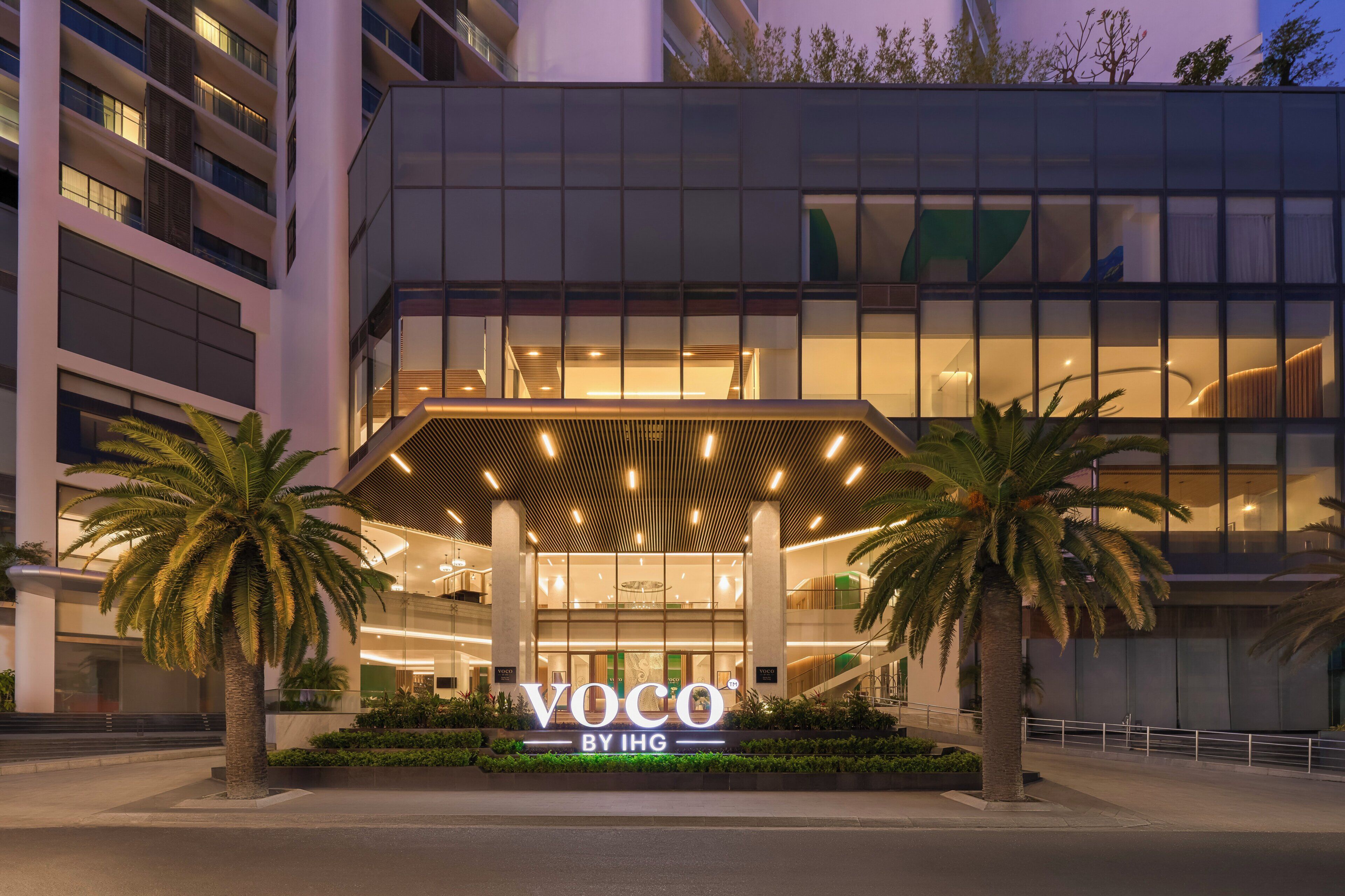 Un edificio moderno con una entrada destacada, palmeras y un cartel que dice 'Voco by IHG'.