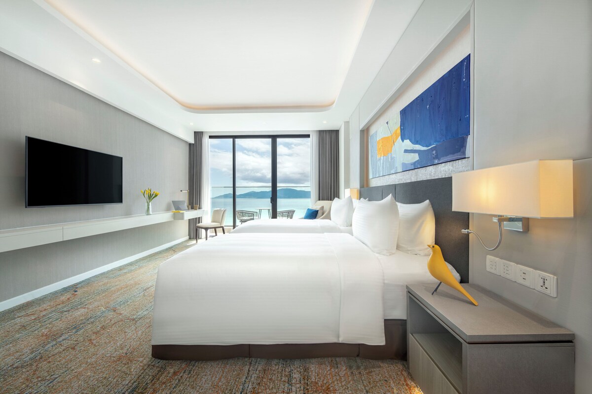 A hotel room with a large bed, a flat-screen TV, a desk with a chair, a painting, and a lamp.