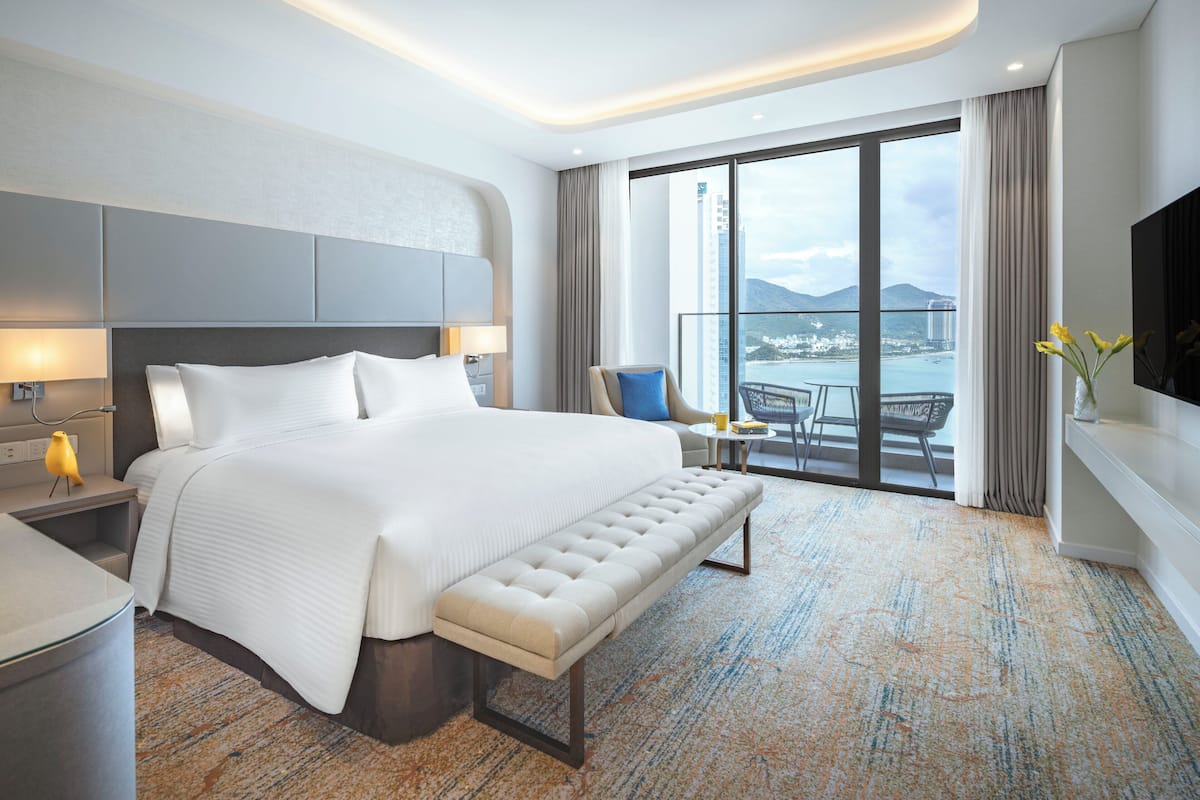 A modern hotel room with a large bed, a bench, a desk, and a view of the city through large windows.