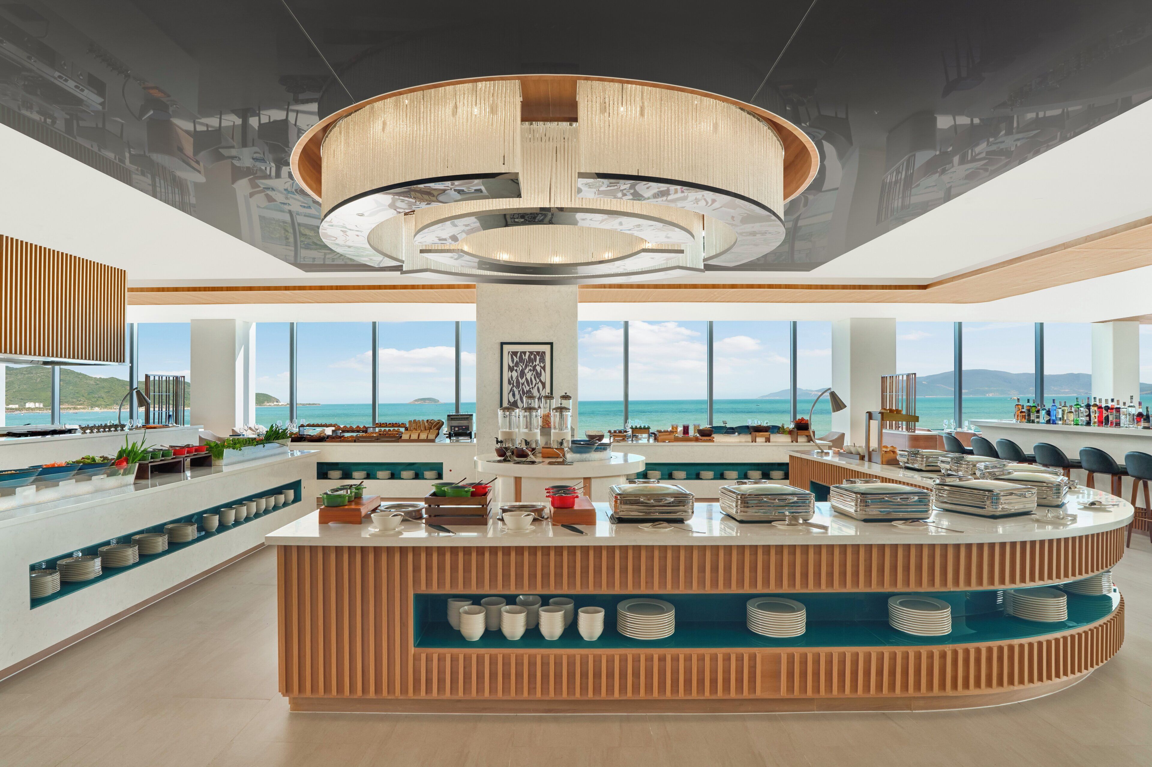 Un hall d’hôtel moderne doté d’un comptoir central, de grandes fenêtres offrant une vue sur la mer et d’un plafond haut orné d’un luminaire circulaire.